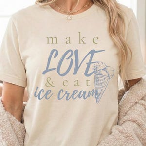 Könnte beinhalten: Cremefarbenes T-Shirt mit dem Text "make LOVE & eat ice cream" in Blau und Grau. Eine blaue Eiswaffel-Grafik ist ebenfalls abgebildet. Das Shirt ist kurzärmelig und aus weichem Material.