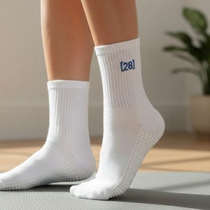Może przedstawiać: Białe skarpetki sportowe z prążkowanym wzorem i antypoślizgowym uchwytem na podeszwach. Skarpetki mają numer "28" wydrukowany na niebiesko u góry. Skarpetki są noszone na szarej macie.