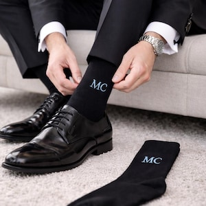 Puede incluir: Una persona con un esmoquin negro se ajusta un calcetín negro con las iniciales "MC" bordadas en azul claro. La persona lleva zapatos de vestir de cuero negro. Una flor blanca y un ojal están prendidos en la solapa del esmoquin.