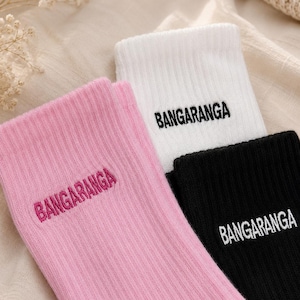 Op de afbeelding: Drie paar geribbelde gebreide sokken in roze, wit en zwart. Elke sok heeft het geborduurde woord "BANGARANGA" in een contrasterende kleur. De sokken zijn gevouwen en gerangschikt op een neutraal gekleurde stof.