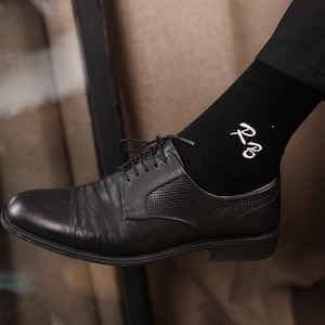 May include: Black leather dress shoe with a cap toe and textured panel. The shoe is paired with a black sock featuring white embroidered initials. The laces are black.
