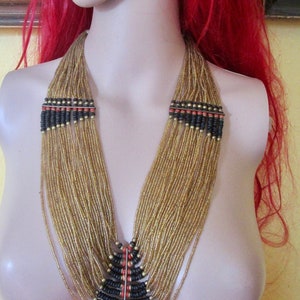 May include: A gold beaded necklace with black and red accents is displayed on a mannequin. The necklace features multiple strands of gold beads, with black and red beaded sections near the top and a triangular design at the bottom.