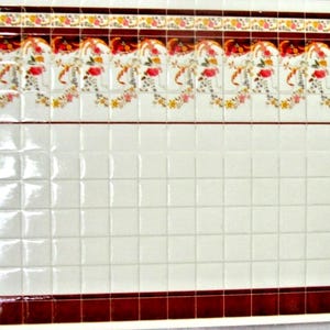 May include: A rectangular grid of glossy white square tiles featuring a decorative border at the top with red, orange, and yellow floral patterns and a solid dark red horizontal stripe along the bottom edge of the tile arrangement.
