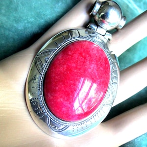 May include: An oval pendant featuring a large, smooth, vibrant red stone set in a polished silver-tone metal frame. The metal border is decorated with intricate engraved patterns, including small lines and dots, and includes a rounded bail.