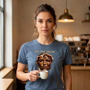 Può includere: Una t-shirt a maniche corte blu mélange con una stampa grafica di un volto formato da vortici di caffè. Sopra la grafica, il testo recita "FIRST COFFEE. THEN EXIST." La maglietta è indossata da una persona che tiene in mano una tazzina bianca.
