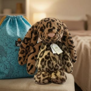May include: A plush leopard-print bunny with long ears and a pink nose sits next to a blue drawstring bag. The bag features a pattern of cartoon characters and the word "Jellycat". The bunny has a tag attached.