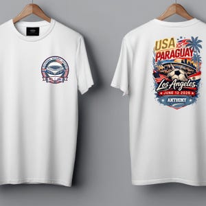 Front and back view of a white t-shirt on hangers featuring a small chest badge on the front and a large colorful soccer graphic on the back reading “USA vs Paraguay, Los Angeles, June 12 2026, Anthony.” The design includes a stadium, soccer ball, palm trees, skyline, and American flag elements. Clean product mockup showcasing a personalized sports fan tee for match day and soccer events.