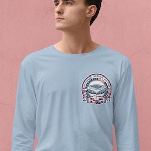 Young man wearing a light blue long sleeve t-shirt with a small chest logo featuring a USA vs Paraguay soccer stadium badge design. Clean minimalist background with a soft pink wall, highlighting a modern and casual look. Comfortable unisex sports fan apparel perfect for soccer lovers, game day outfits, and everyday wear.