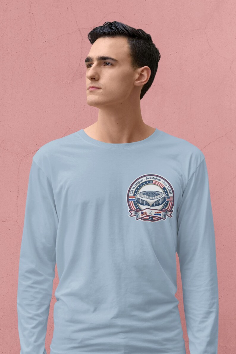 Young man wearing a light blue long sleeve t-shirt with a small chest logo featuring a USA vs Paraguay soccer stadium badge design. Clean minimalist background with a soft pink wall, highlighting a modern and casual look. Comfortable unisex sports fan apparel perfect for soccer lovers, game day outfits, and everyday wear.