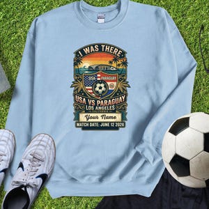 Light blue sweatshirt mockup featuring a personalized vintage-style “I Was There” USA vs Paraguay soccer match design. Graphic includes a sunset stadium scene, palm trees, soccer ball, USA and Paraguay flags, “Los Angeles California,” customizable name text, and “Match Date: June 12 2026.” Displayed flat lay on green grass with soccer cleats and a soccer ball, perfect personalized souvenir sweatshirt for soccer fans, match day, and sports memories.