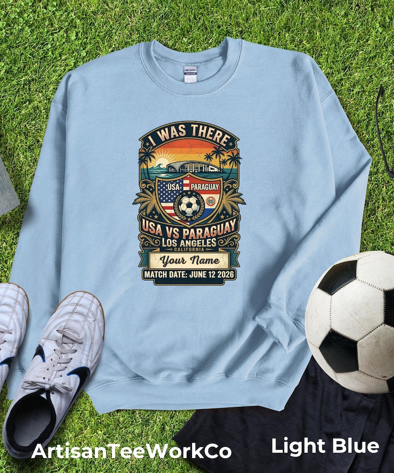 Light blue sweatshirt mockup featuring a personalized vintage-style “I Was There” USA vs Paraguay soccer match design. Graphic includes a sunset stadium scene, palm trees, soccer ball, USA and Paraguay flags, “Los Angeles California,” customizable name text, and “Match Date: June 12 2026.” Displayed flat lay on green grass with soccer cleats and a soccer ball, perfect personalized souvenir sweatshirt for soccer fans, match day, and sports memories.