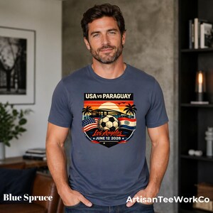 Man wearing a blue spruce t-shirt indoors in a modern home setting, featuring a bold vintage-style soccer match design reading “USA vs Paraguay,” “Los Angeles,” and “June 12 2028.” Artwork includes a soccer ball, stadium silhouette, sunset background, palm trees, and USA and Paraguay flags, making it a stylish commemorative soccer fan shirt perfect for match day, travel, or patriotic sportswear.