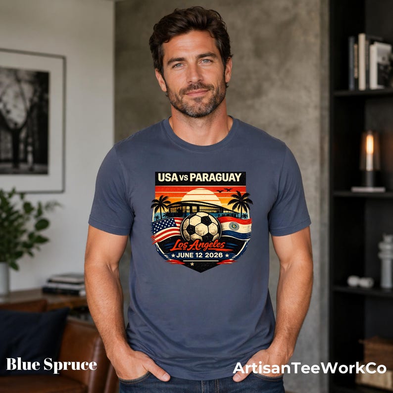Man wearing a blue spruce t-shirt indoors in a modern home setting, featuring a bold vintage-style soccer match design reading “USA vs Paraguay,” “Los Angeles,” and “June 12 2028.” Artwork includes a soccer ball, stadium silhouette, sunset background, palm trees, and USA and Paraguay flags, making it a stylish commemorative soccer fan shirt perfect for match day, travel, or patriotic sportswear.