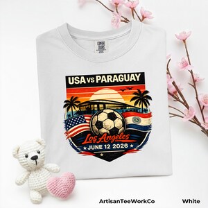 Folded white Comfort Colors t-shirt displayed with pink floral accents and a teddy bear prop, featuring a bold vintage-style soccer match design reading “USA vs Paraguay,” “Los Angeles,” and “June 12 2028.” Artwork includes a soccer ball, stadium silhouette, sunset background, palm trees, and USA and Paraguay flags, making it a stylish commemorative soccer fan shirt for match day, travel, or patriotic sportswear.