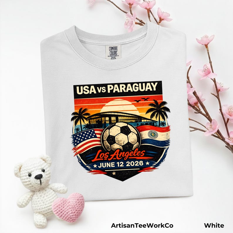Folded white Comfort Colors t-shirt displayed with pink floral accents and a teddy bear prop, featuring a bold vintage-style soccer match design reading “USA vs Paraguay,” “Los Angeles,” and “June 12 2028.” Artwork includes a soccer ball, stadium silhouette, sunset background, palm trees, and USA and Paraguay flags, making it a stylish commemorative soccer fan shirt for match day, travel, or patriotic sportswear.