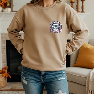 Woman wearing a beige crewneck sweatshirt with a small chest logo featuring a USA vs Paraguay soccer stadium badge design. Styled with blue ripped jeans in a cozy home setting with fireplace and autumn decor. Casual and comfortable sports fan apparel perfect for soccer lovers, game day outfits, and everyday wear.
