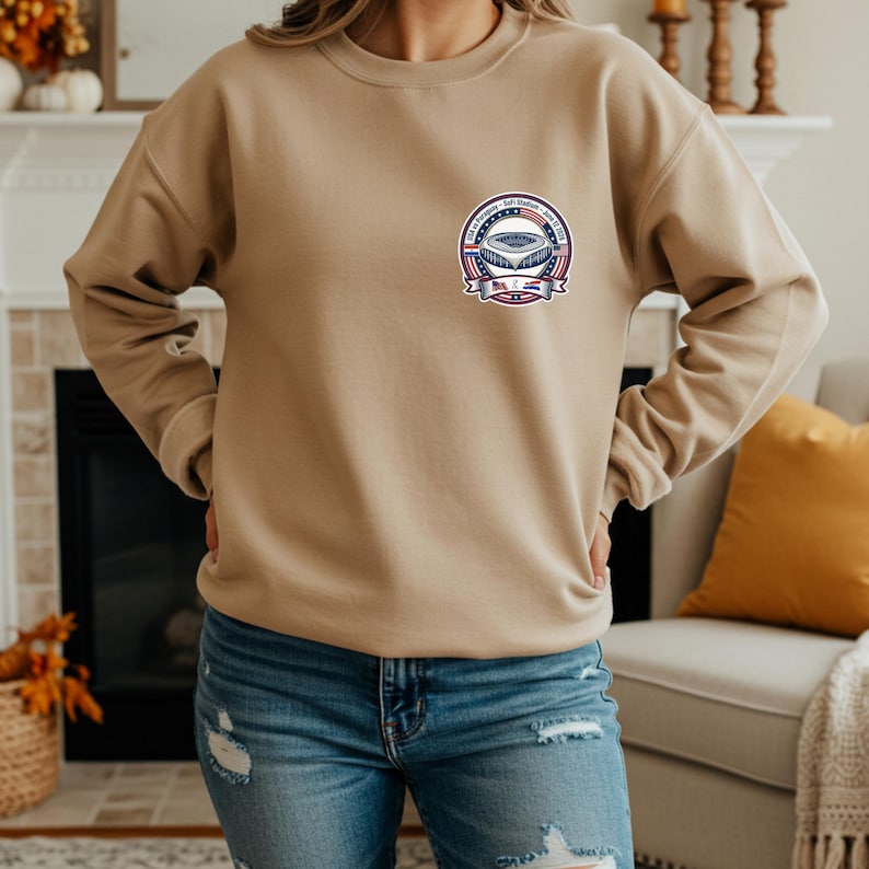 Woman wearing a beige crewneck sweatshirt with a small chest logo featuring a USA vs Paraguay soccer stadium badge design. Styled with blue ripped jeans in a cozy home setting with fireplace and autumn decor. Casual and comfortable sports fan apparel perfect for soccer lovers, game day outfits, and everyday wear.