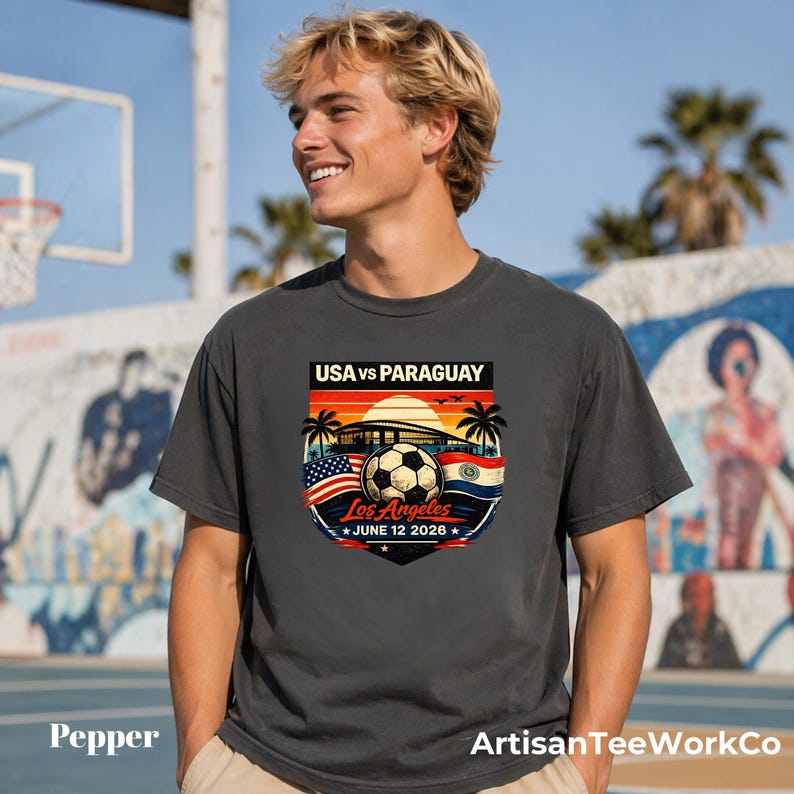 Young man wearing a pepper dark gray t-shirt outdoors on a sunny basketball court, featuring a bold vintage-style soccer match design reading “USA vs Paraguay,” “Los Angeles,” and “June 12 2028.” Artwork includes a soccer ball, stadium silhouette, sunset background, palm trees, and USA and Paraguay flags, making it a stylish commemorative soccer fan shirt perfect for match day, travel, or patriotic sportswear.