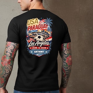 Back view of a man wearing a black t-shirt with a bold soccer match graphic reading “USA vs Paraguay, Los Angeles, June 12 2026, Anthony.” The design features a stadium, soccer ball, palm trees, and American flag elements in vibrant red, white, and blue. Lifestyle mockup highlighting a sports fan graphic tee for soccer lovers and match day events.