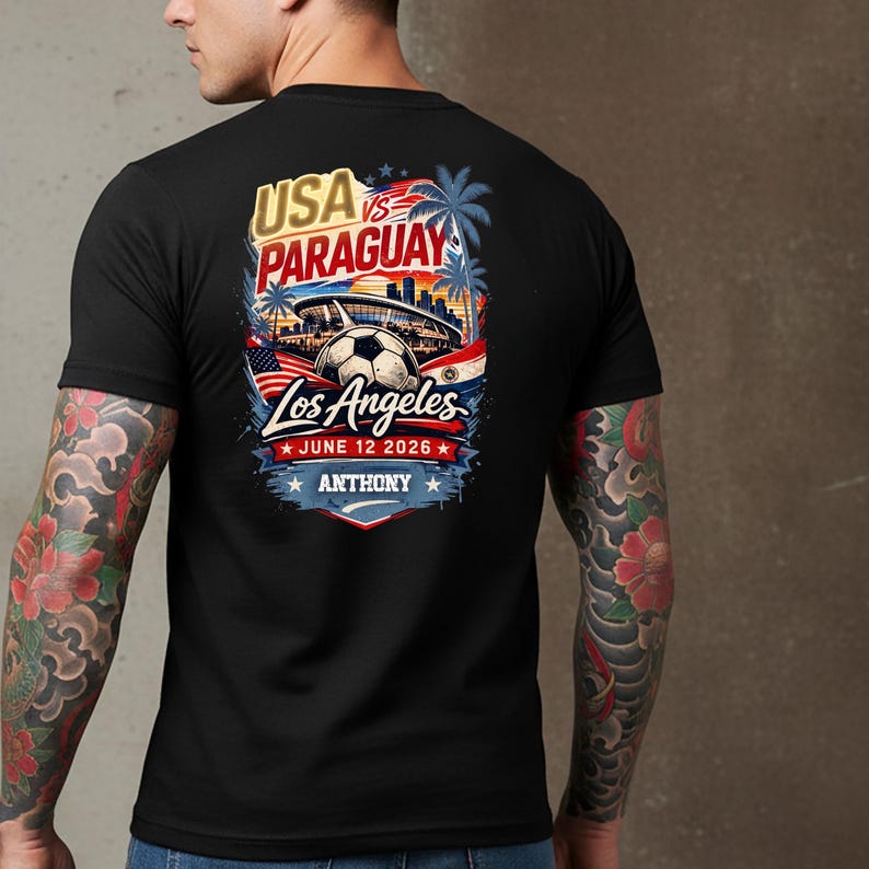 Back view of a man wearing a black t-shirt with a bold soccer match graphic reading “USA vs Paraguay, Los Angeles, June 12 2026, Anthony.” The design features a stadium, soccer ball, palm trees, and American flag elements in vibrant red, white, and blue. Lifestyle mockup highlighting a sports fan graphic tee for soccer lovers and match day events.