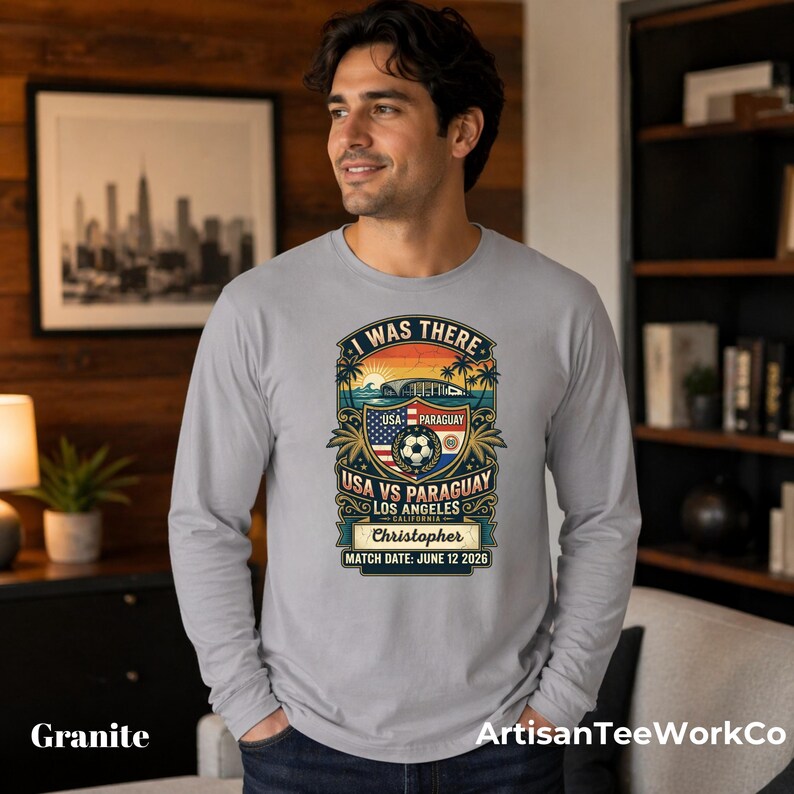 Men’s granite gray long sleeve t-shirt mockup featuring a personalized vintage-style “I Was There” USA vs Paraguay soccer match design. Graphic includes a sunset stadium scene, palm trees, soccer ball, USA and Paraguay flags, “Los Angeles California,” custom name “Christopher,” and “Match Date: June 12 2026.” Modeled by a man indoors in a modern living room setting. Perfect personalized soccer fan souvenir shirt for match day and sports memories.
