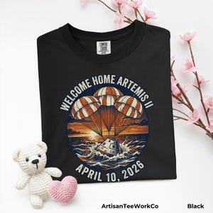 May include: Black t-shirt with a graphic design featuring the text "WELCOME HOME ARTEMIS II" and the date "APRIL 10, 2026". The design includes parachutes, a spacecraft, and ocean waves. The shirt has a crew neck.