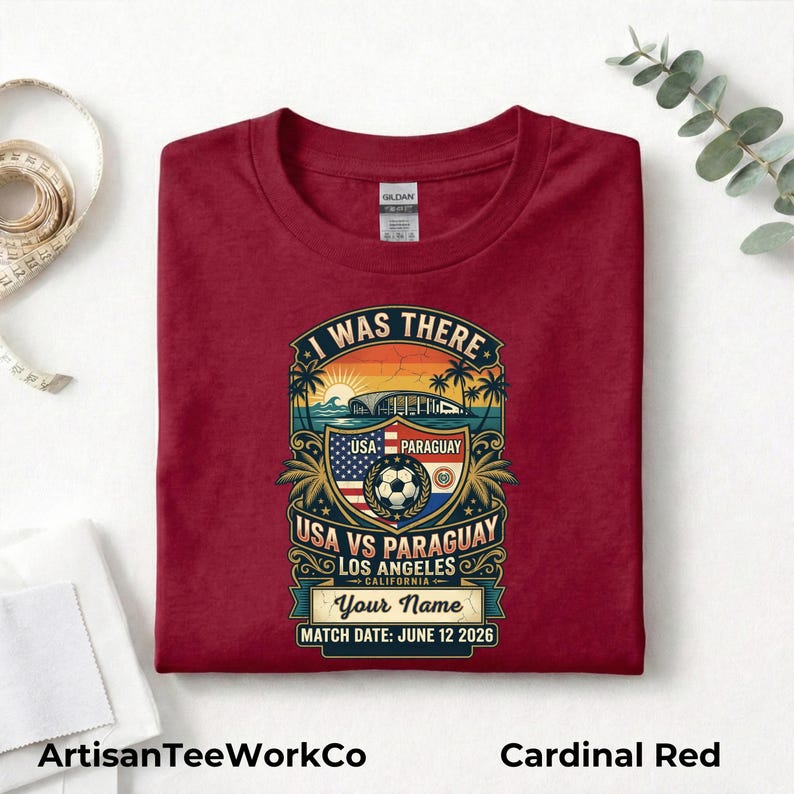 Folded cardinal red t-shirt mockup featuring a personalized vintage-style soccer match design with “I Was There,” “USA vs Paraguay,” “Los Angeles California,” customizable name text, and “Match Date: June 12 2026.” The retro graphic includes a soccer ball, stadium silhouette, sunset, palm trees, and USA and Paraguay flags. Displayed on a clean white background with measuring tape and eucalyptus accents. Perfect keepsake shirt for soccer fans, match day, or commemorative event apparel.