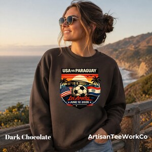 Smiling woman wearing a dark chocolate brown sweatshirt outdoors by a scenic ocean coastline at sunset, featuring a bold vintage-style soccer match graphic that reads “USA vs Paraguay,” “Los Angeles,” and “June 12 2028.” The colorful design includes a soccer ball, stadium silhouette, sunset, palm trees, and USA and Paraguay flags. Perfect for soccer fans, match day outfits, travel, sports events, or patriotic game day apparel.
