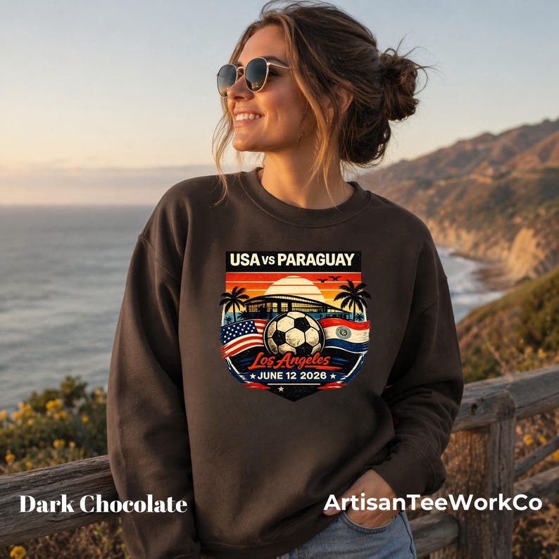 Smiling woman wearing a dark chocolate brown sweatshirt outdoors by a scenic ocean coastline at sunset, featuring a bold vintage-style soccer match graphic that reads “USA vs Paraguay,” “Los Angeles,” and “June 12 2028.” The colorful design includes a soccer ball, stadium silhouette, sunset, palm trees, and USA and Paraguay flags. Perfect for soccer fans, match day outfits, travel, sports events, or patriotic game day apparel.