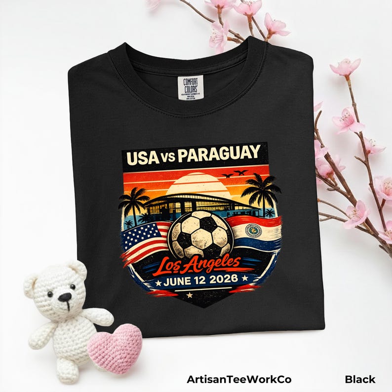 Folded black Comfort Colors t-shirt displayed with pink floral accents and a teddy bear prop, featuring a bold vintage-style soccer match design reading “USA vs Paraguay,” “Los Angeles,” and “June 12 2026.” Artwork includes a soccer ball, stadium silhouette, sunset background, palm trees, and USA and Paraguay flags, making it a stylish commemorative soccer fan shirt for match day or patriotic sportswear.