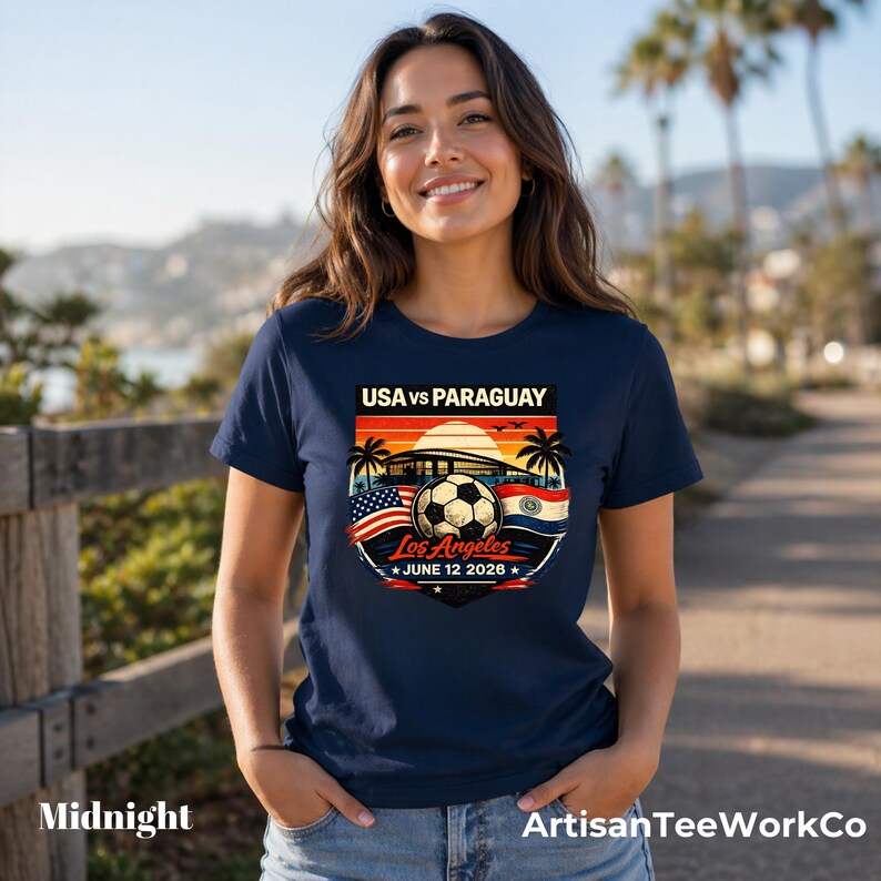 Woman wearing a midnight navy t-shirt while walking outdoors on a sunny palm-lined path, featuring a bold vintage-style soccer match design reading “USA vs Paraguay,” “Los Angeles,” and “June 12 2028.” Artwork includes a soccer ball, stadium silhouette, sunset background, palm trees, and USA and Paraguay flags, making it a stylish commemorative soccer fan shirt perfect for match day, travel, or patriotic sportswear.