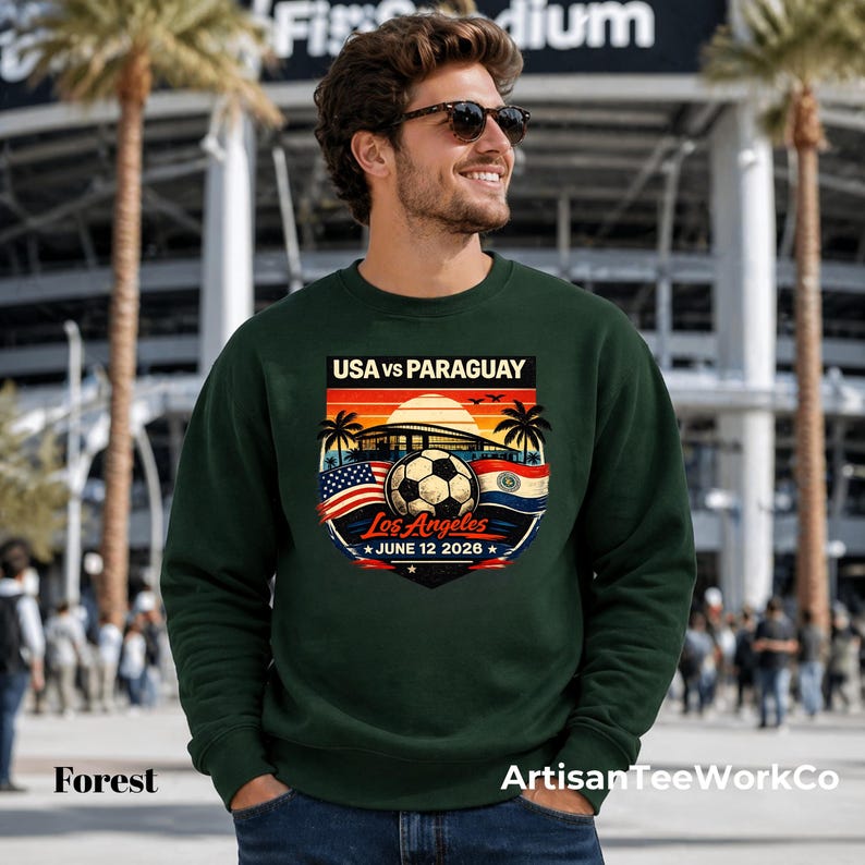 Smiling man wearing a forest green sweatshirt outside a stadium, featuring a bold vintage-style soccer match graphic that reads “USA vs Paraguay,” “Los Angeles,” and “June 12 2028.” The colorful retro design includes a soccer ball, stadium silhouette, sunset, palm trees, and USA and Paraguay flags. Perfect for soccer fans, match day outfits, sports events, stadium visits, travel, or patriotic game day apparel.