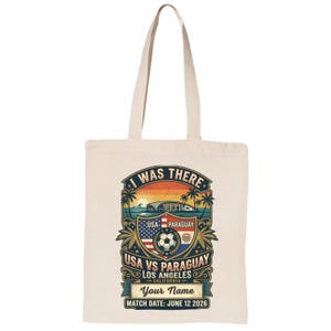 Natural canvas tote bag featuring a personalized vintage-style “I Was There” USA vs Paraguay soccer match design. Graphic includes a sunset stadium scene, palm trees, soccer ball, USA and Paraguay flags, “Los Angeles California,” customizable name text, and “Match Date: June 12 2026.” Reusable eco-friendly tote bag mockup on a plain light background, perfect personalized soccer fan gift, match day souvenir, and everyday carryall bag.