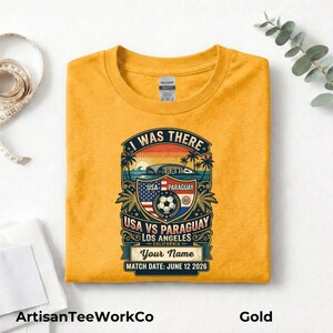 Folded gold t-shirt mockup featuring a personalized vintage-style soccer match design with “I Was There,” “USA vs Paraguay,” “Los Angeles California,” customizable name text, and “Match Date: June 12 2026.” The retro graphic includes a soccer ball, stadium silhouette, sunset, palm trees, and USA and Paraguay flags in bold distressed colors. Displayed on a clean white background with measuring tape, folded fabric, and eucalyptus accents.