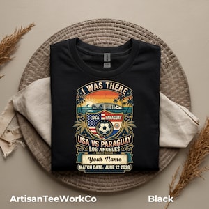Black folded t-shirt mockup featuring a personalized vintage-style soccer match design with “I Was There,” “USA vs Paraguay,” “Los Angeles California,” customizable name text, and “Match Date: June 12 2026.” The retro graphic includes a soccer ball, stadium silhouette, sunset, palm trees, and USA and Paraguay flags. Displayed on a woven round mat with neutral decor accents. Perfect keepsake shirt for soccer fans, match day, or commemorative event apparel.