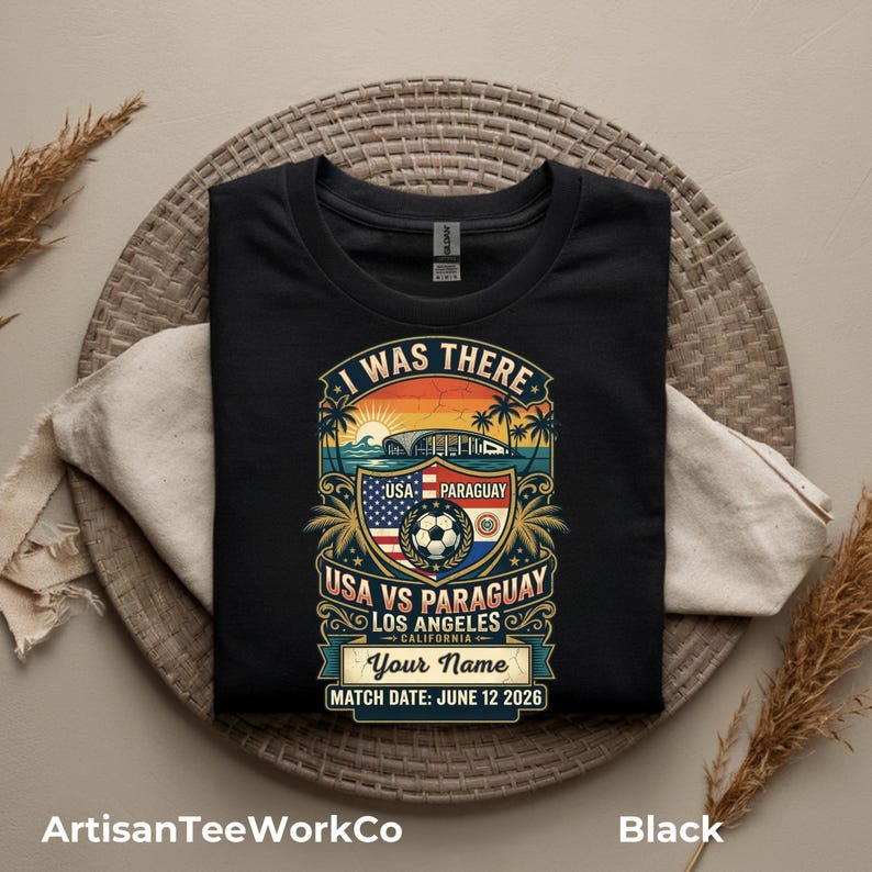 Black folded t-shirt mockup featuring a personalized vintage-style soccer match design with “I Was There,” “USA vs Paraguay,” “Los Angeles California,” customizable name text, and “Match Date: June 12 2026.” The retro graphic includes a soccer ball, stadium silhouette, sunset, palm trees, and USA and Paraguay flags. Displayed on a woven round mat with neutral decor accents. Perfect keepsake shirt for soccer fans, match day, or commemorative event apparel.
