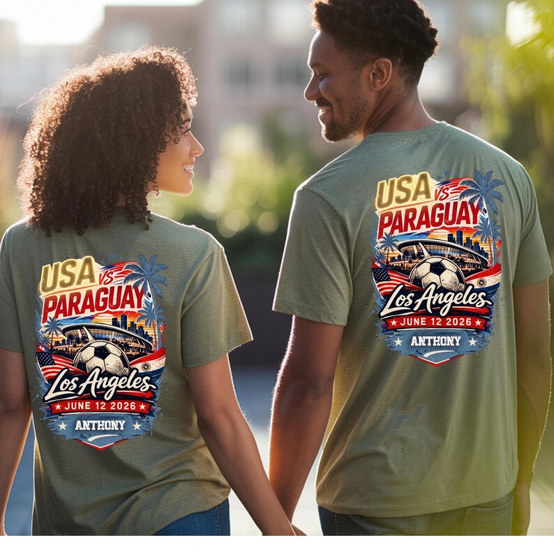 Back view of a couple wearing matching olive green t-shirts featuring a bold USA vs Paraguay soccer graphic with Los Angeles skyline, stadium, palm trees, and soccer ball, dated June 12 2026 with personalized name Anthony. Walking outdoors in sunlight, showcasing stylish unisex sports fan apparel perfect for soccer lovers, game day outfits, and casual wear.
