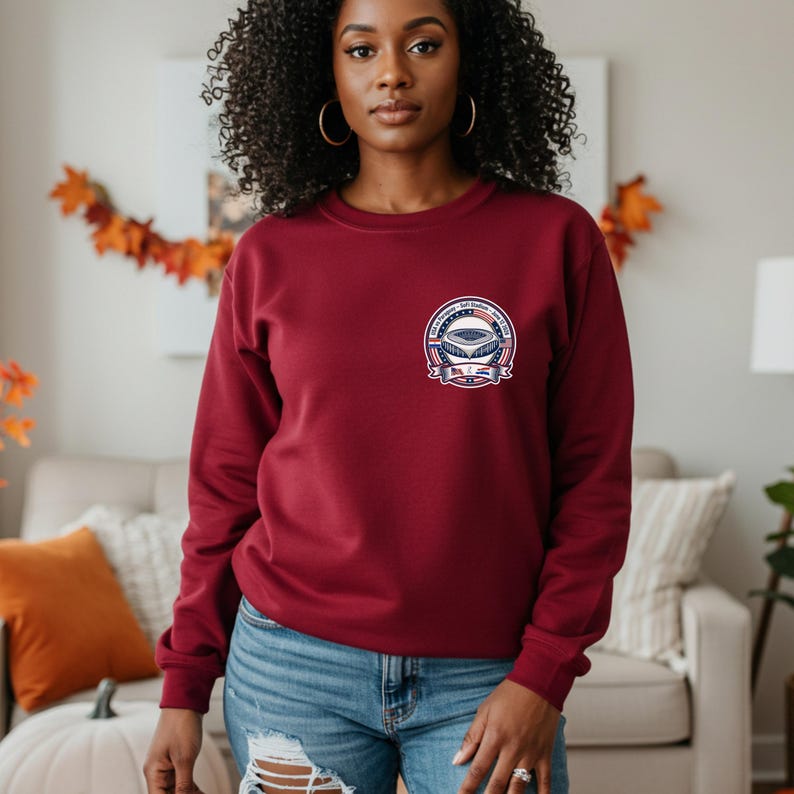 Woman wearing a burgundy crewneck sweatshirt with a small chest graphic featuring a soccer stadium badge design inspired by USA vs Paraguay match. Casual fall setting with cozy home decor and autumn leaves in the background. Stylish outfit paired with jeans, showcasing a comfortable sports fan sweatshirt perfect for game day, soccer lovers, and everyday wear.