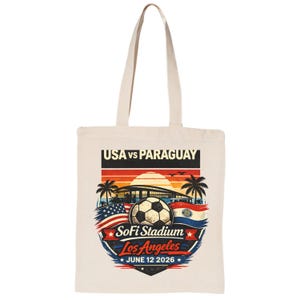 May include: Cream-colored tote bag featuring a soccer ball and the text "USA vs Paraguay" above a stadium graphic. The design includes the flags of the USA and Paraguay, palm trees, and the text "SoFi Stadium Los Angeles June 12 2026."