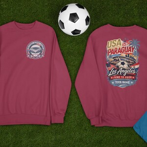Two maroon sweatshirts displayed on green grass with a soccer ball and referee whistle nearby, showing front and back designs. Front features a small USA vs Paraguay soccer badge with stadium graphic, while back showcases a large vibrant Los Angeles match design with palm trees, stadium, soccer ball, and date June 12 2026 with customizable name text. Casual sports fan apparel perfect for soccer lovers and game day outfits.