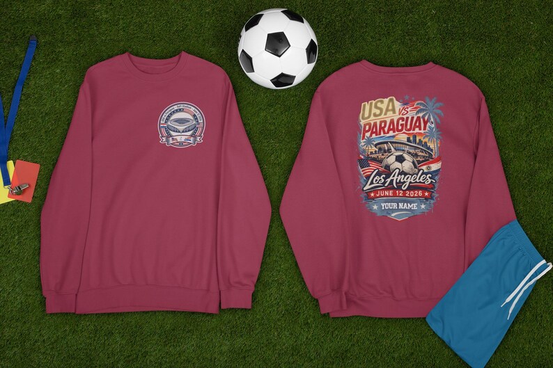 Two maroon sweatshirts displayed on green grass with a soccer ball and referee whistle nearby, showing front and back designs. Front features a small USA vs Paraguay soccer badge with stadium graphic, while back showcases a large vibrant Los Angeles match design with palm trees, stadium, soccer ball, and date June 12 2026 with customizable name text. Casual sports fan apparel perfect for soccer lovers and game day outfits.