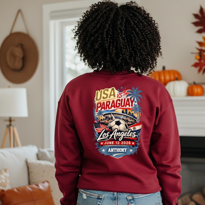 Back view of a woman wearing a burgundy sweatshirt featuring a bold USA vs Paraguay soccer graphic with Los Angeles skyline, stadium, palm trees, and soccer ball, dated June 12 2026 with personalized name Anthony. Cozy indoor fall setting with pumpkins and warm decor, highlighting a stylish sports fan sweatshirt perfect for soccer lovers and game day apparel.