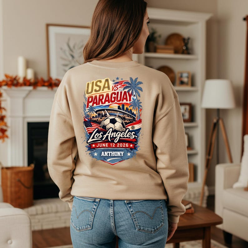 Back view of a woman wearing a beige sweatshirt featuring a vibrant USA vs Paraguay soccer graphic with Los Angeles skyline, stadium, palm trees, and soccer ball, dated June 12 2026 with personalized name Anthony. Styled with blue jeans in a cozy home setting with fireplace and fall decor, perfect for soccer fans, game day outfits, and casual everyday wear.