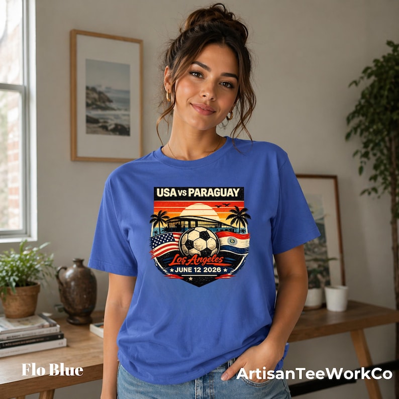 Woman wearing a flo blue t-shirt indoors in a cozy home setting, featuring a bold vintage-style soccer match design reading “USA vs Paraguay,” “Los Angeles,” and “June 12 2028.” Artwork includes a soccer ball, stadium silhouette, sunset background, palm trees, and USA and Paraguay flags, making it a stylish commemorative soccer fan shirt perfect for match day, travel, or patriotic sportswear.