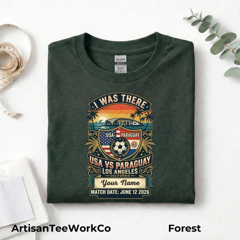 Folded forest green t-shirt mockup featuring a personalized vintage-style soccer match design with “I Was There,” “USA vs Paraguay,” “Los Angeles California,” customizable name text, and “Match Date: June 12 2026.” The retro graphic includes a soccer ball, stadium silhouette, sunset, palm trees, and USA and Paraguay flags. Displayed on a clean white background with measuring tape and eucalyptus accents. Perfect keepsake shirt for soccer fans, match day, or commemorative event apparel.
