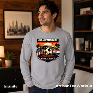 Smiling man wearing a granite gray long sleeve shirt indoors in a cozy modern room, featuring a bold vintage-style soccer match graphic that reads “USA vs Paraguay,” “Los Angeles,” and “June 12 2028.” The colorful design includes a soccer ball, stadium silhouette, sunset, palm trees, and USA and Paraguay flags. Great for soccer fans, match day outfits, sports events, travel, or patriotic game day apparel.