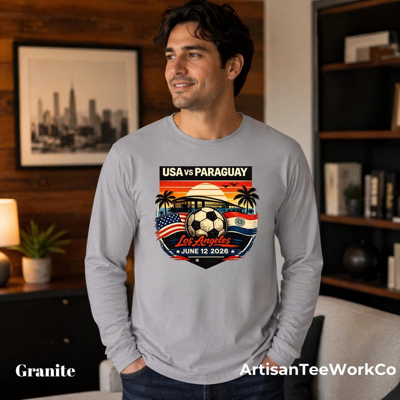 Smiling man wearing a granite gray long sleeve shirt indoors in a cozy modern room, featuring a bold vintage-style soccer match graphic that reads “USA vs Paraguay,” “Los Angeles,” and “June 12 2028.” The colorful design includes a soccer ball, stadium silhouette, sunset, palm trees, and USA and Paraguay flags. Great for soccer fans, match day outfits, sports events, travel, or patriotic game day apparel.