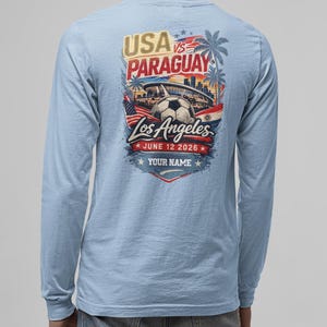 Back view of a man wearing a light blue long sleeve t-shirt featuring a bold USA vs Paraguay soccer graphic with Los Angeles skyline, stadium, palm trees, and soccer ball, dated June 12 2026 with customizable text “Your Name.” Casual denim styling on a clean background, highlighting comfortable unisex sports fan apparel perfect for soccer lovers and game day wear.