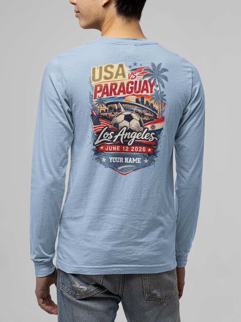 Back view of a man wearing a light blue long sleeve t-shirt featuring a bold USA vs Paraguay soccer graphic with Los Angeles skyline, stadium, palm trees, and soccer ball, dated June 12 2026 with customizable text “Your Name.” Casual denim styling on a clean background, highlighting comfortable unisex sports fan apparel perfect for soccer lovers and game day wear.