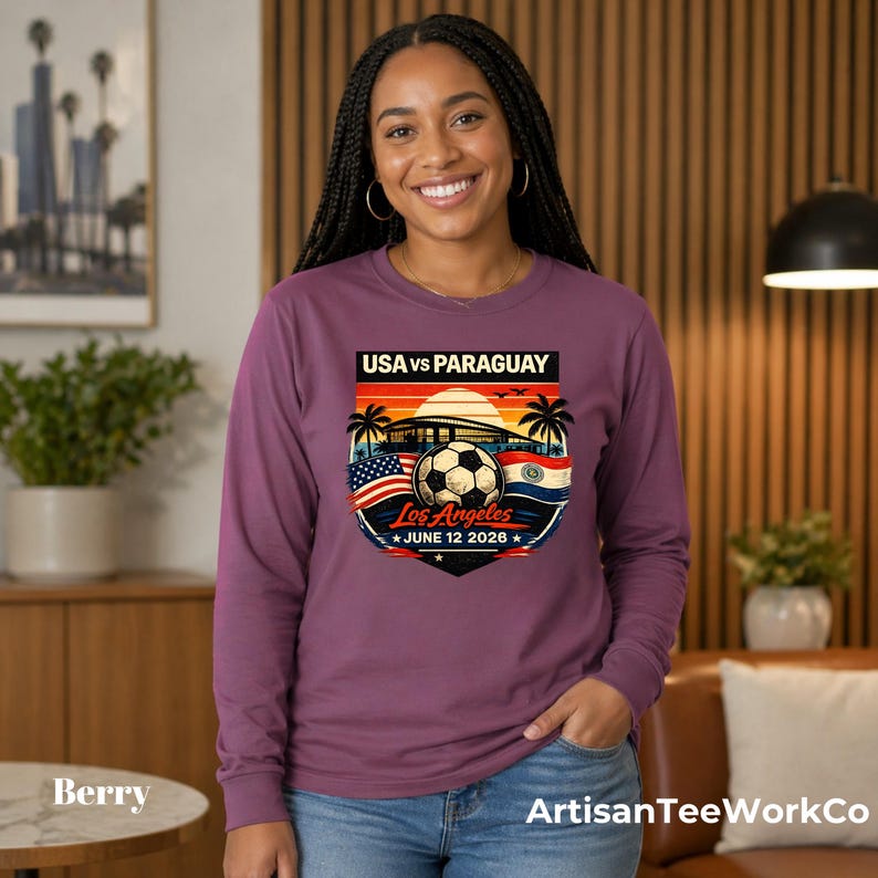 Smiling woman wearing a berry long sleeve shirt indoors in a modern home setting, featuring a bold vintage-style soccer match graphic that reads “USA vs Paraguay,” “Los Angeles,” and “June 12 2028.” The colorful design includes a soccer ball, stadium silhouette, sunset, palm trees, and USA and Paraguay flags. Perfect for soccer fans, match day outfits, sports events, travel, or patriotic game day apparel.
